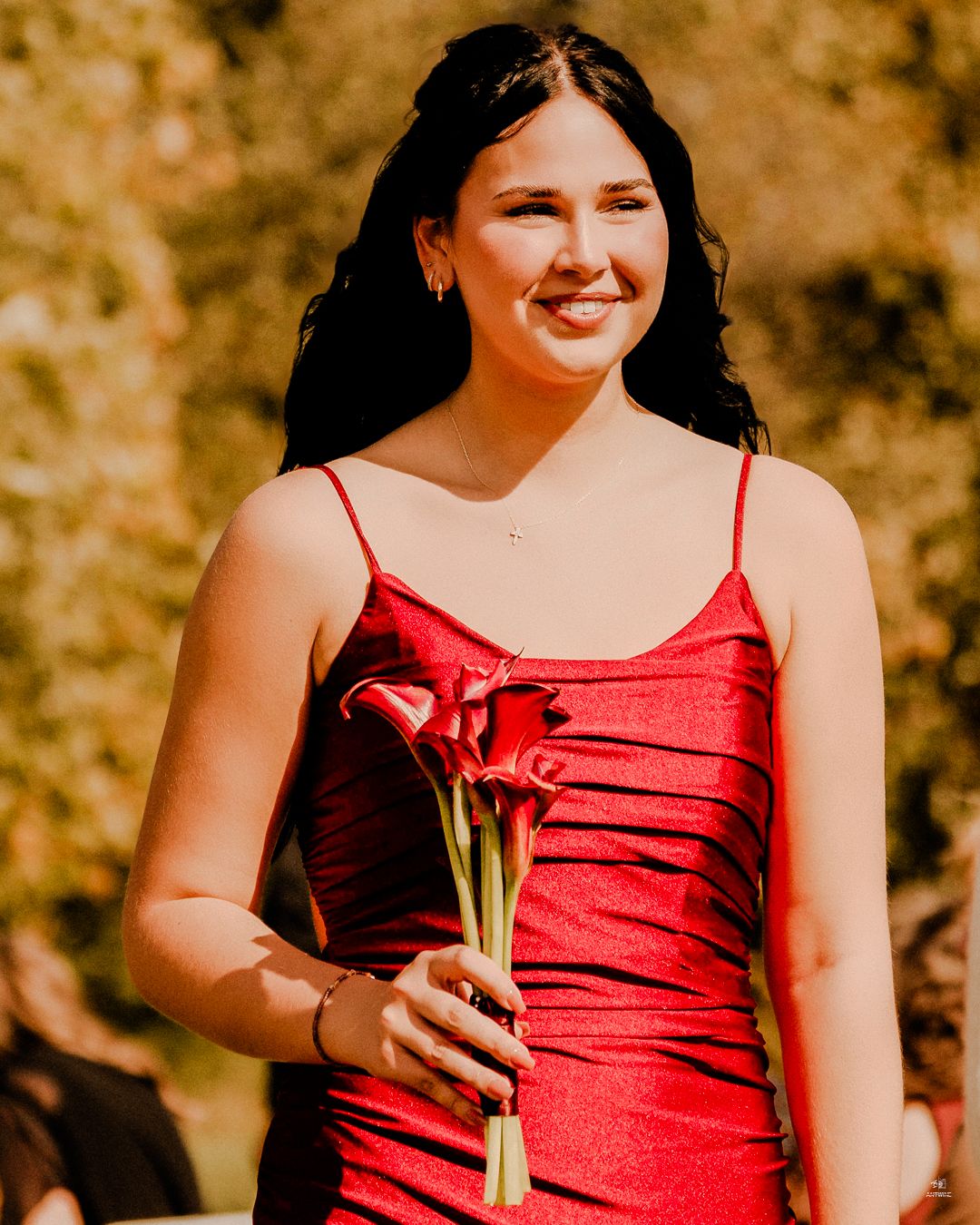 Smiling bridesmaid in red spaghetti strap dress holding red calla lily flowers at outdoor wedding.