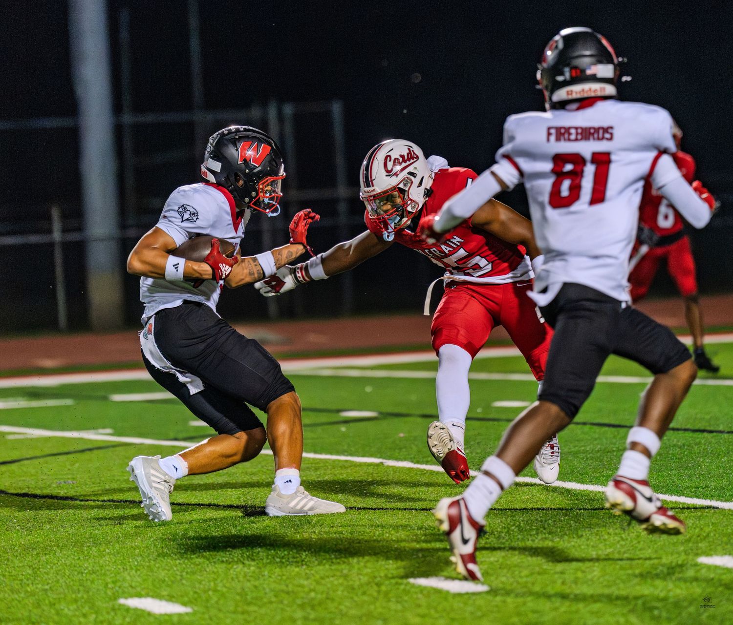 Football player in dark uniform stiff-arms red-jersied defender during high school night game action.