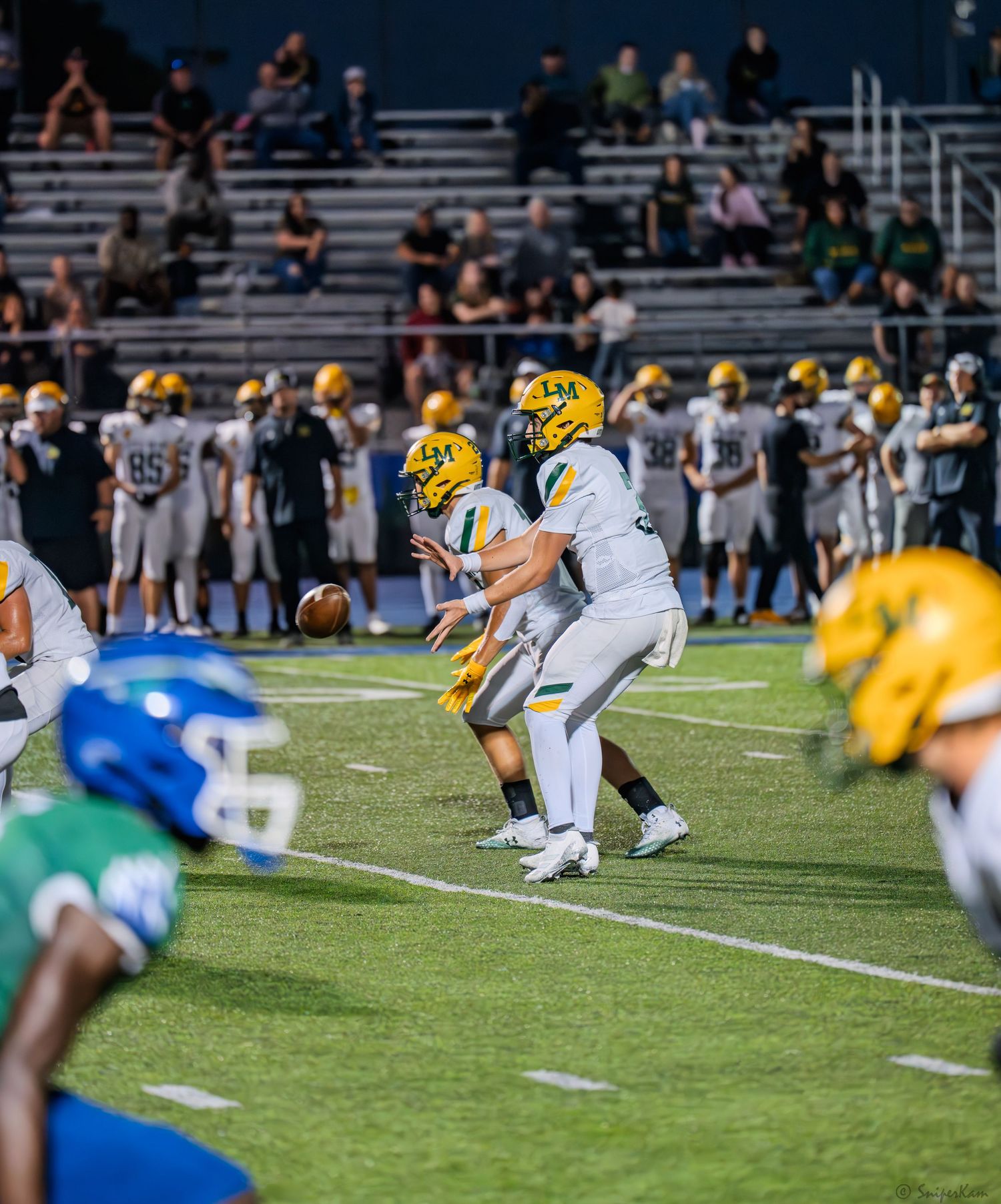 High school football players in gold helmets line up at scrimmage against blue and green opponents under stadium lights.