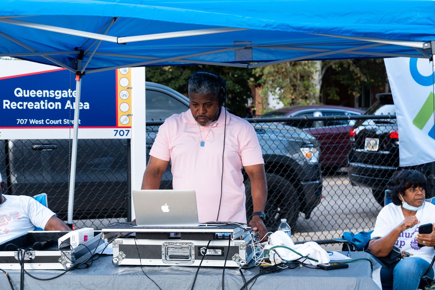 DJ performs at Queensgate Recreation Area outdoor community event, operating laptop and audio equipment under blue canopy tent.