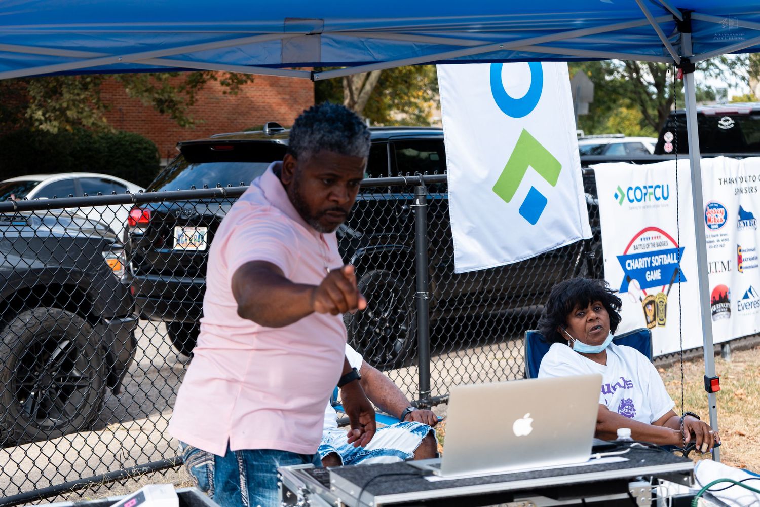 DJ performing at outdoor community event under blue tent with laptop and equipment, banner visible in background.