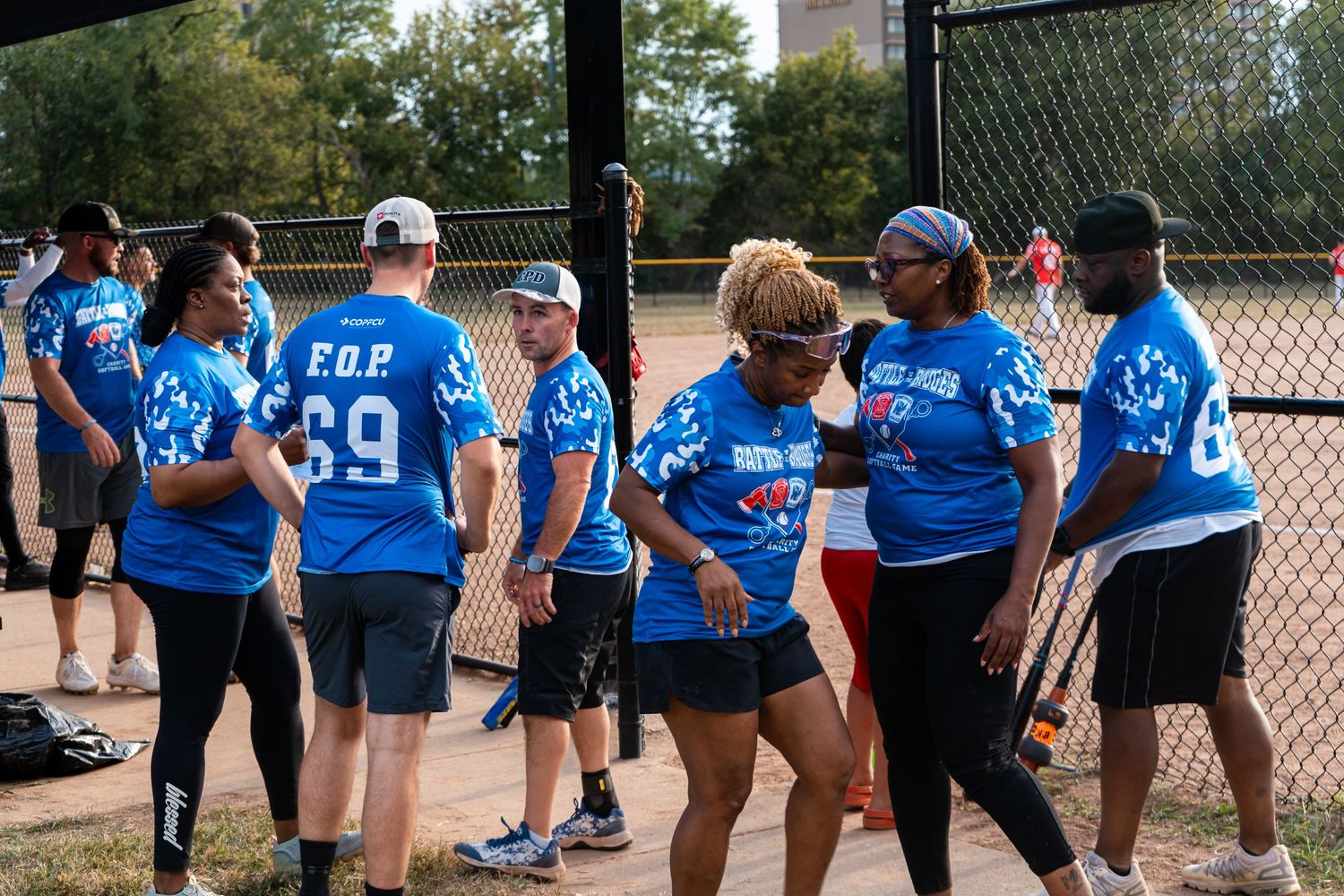 Players in blue FOP jerseys gather and interact on a softball field during a charity game event.
