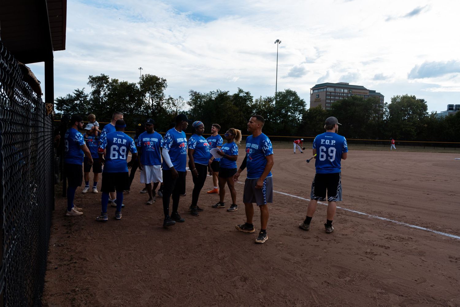 A team in blue jerseys numbered 69 gathers on a dirt softball field during an evening game.