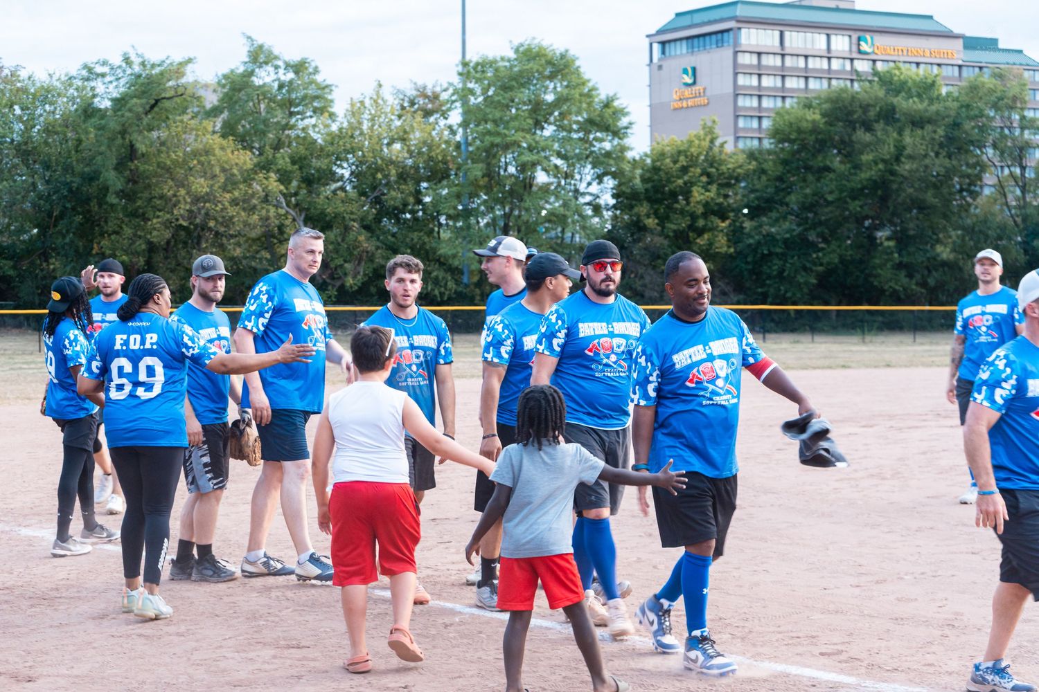 Blue team softball players high-five children on a dirt baseball field during a community game outdoors.