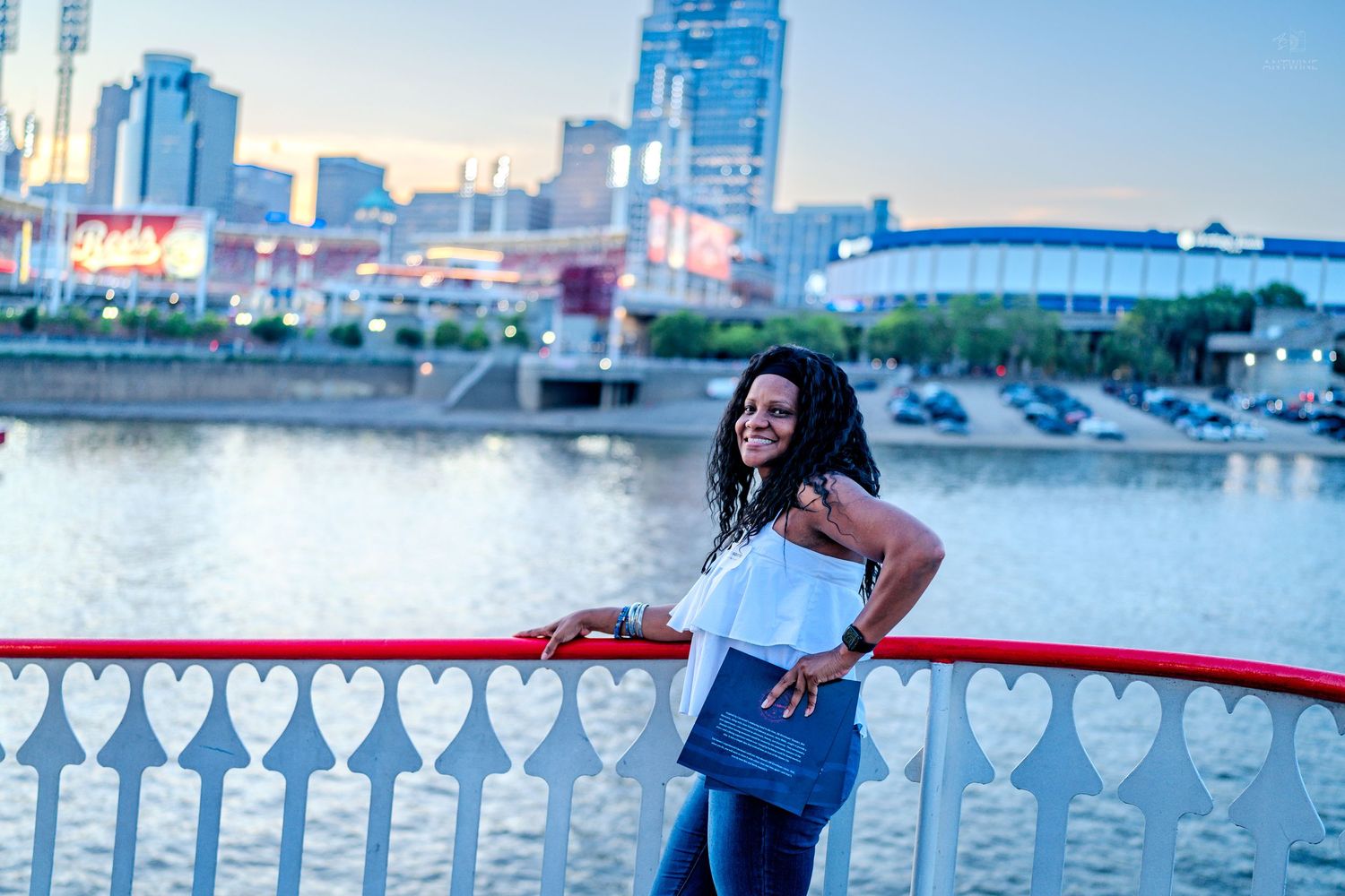 Woman smiling on a red railing bridge over a river with a city skyline at dusk behind her.