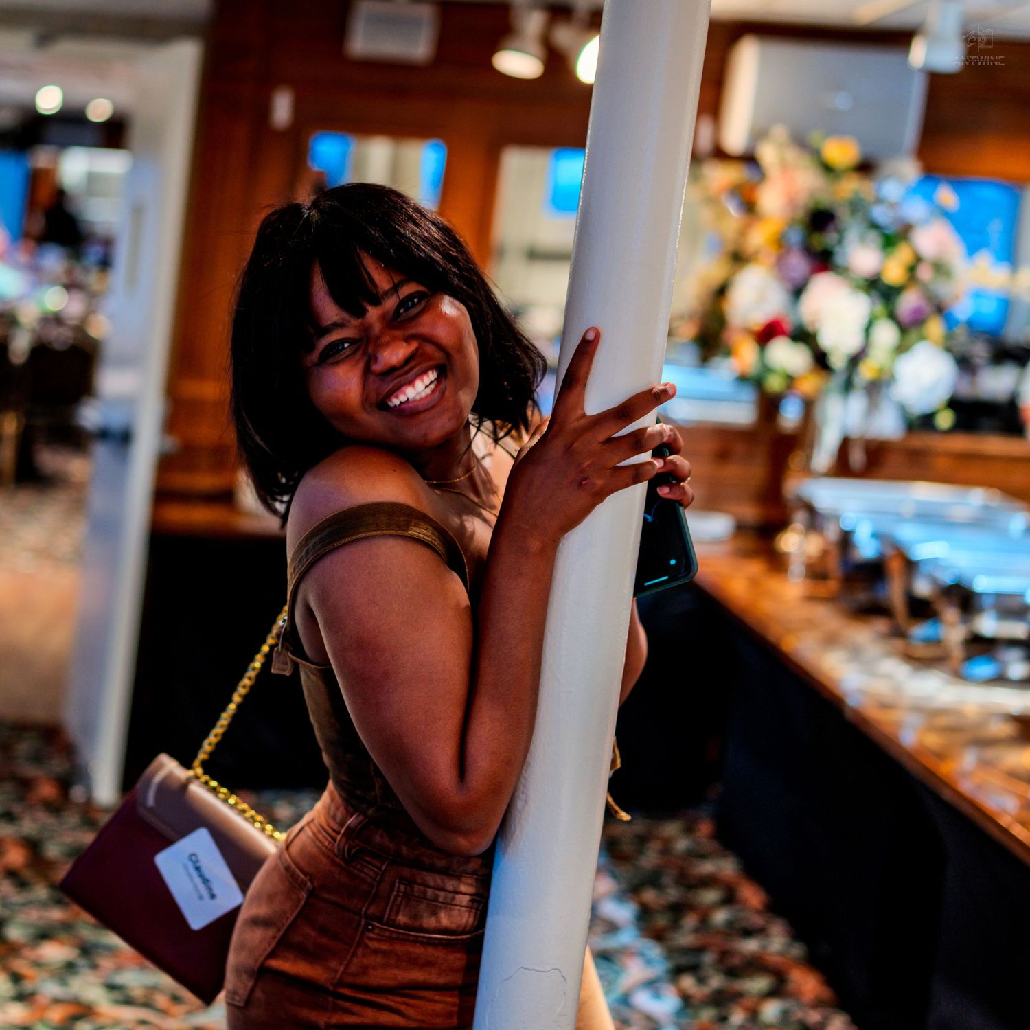 Smiling woman in brown outfit holding a bottle, leaning against a white pillar in an elegant restaurant.