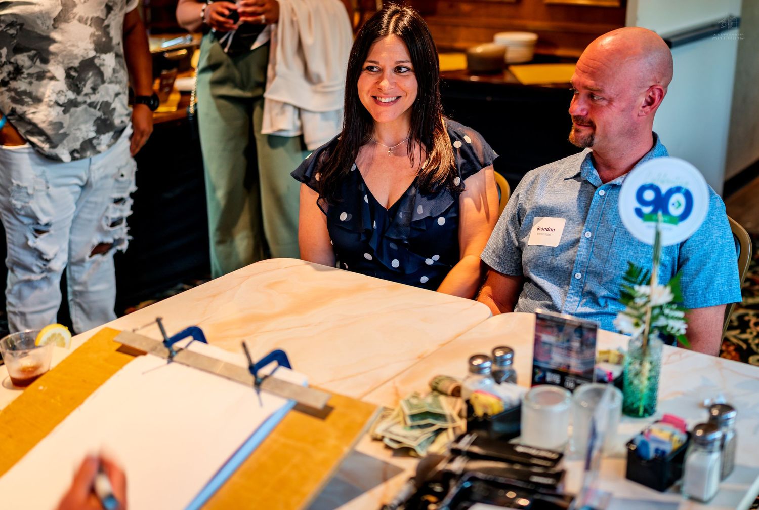 Smiling woman and bald man sitting at a caricature artist table at a 90th birthday celebration.