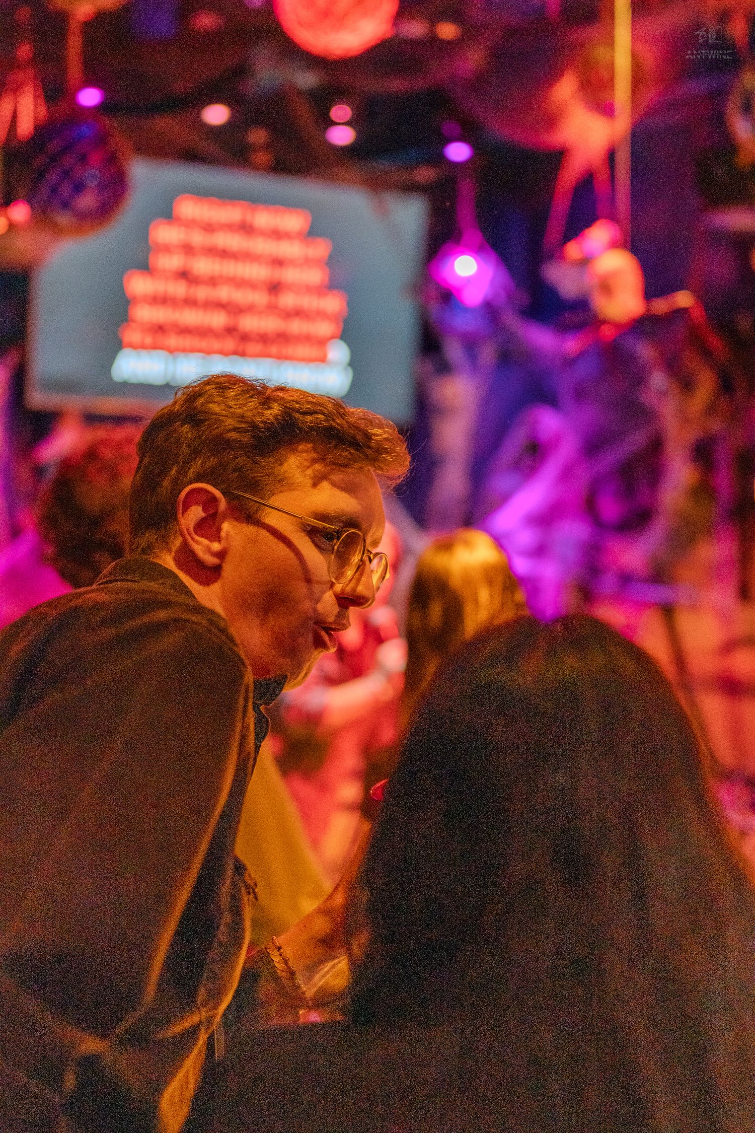 Man with glasses at dimly lit Halloween karaoke bar with purple and orange neon lighting.