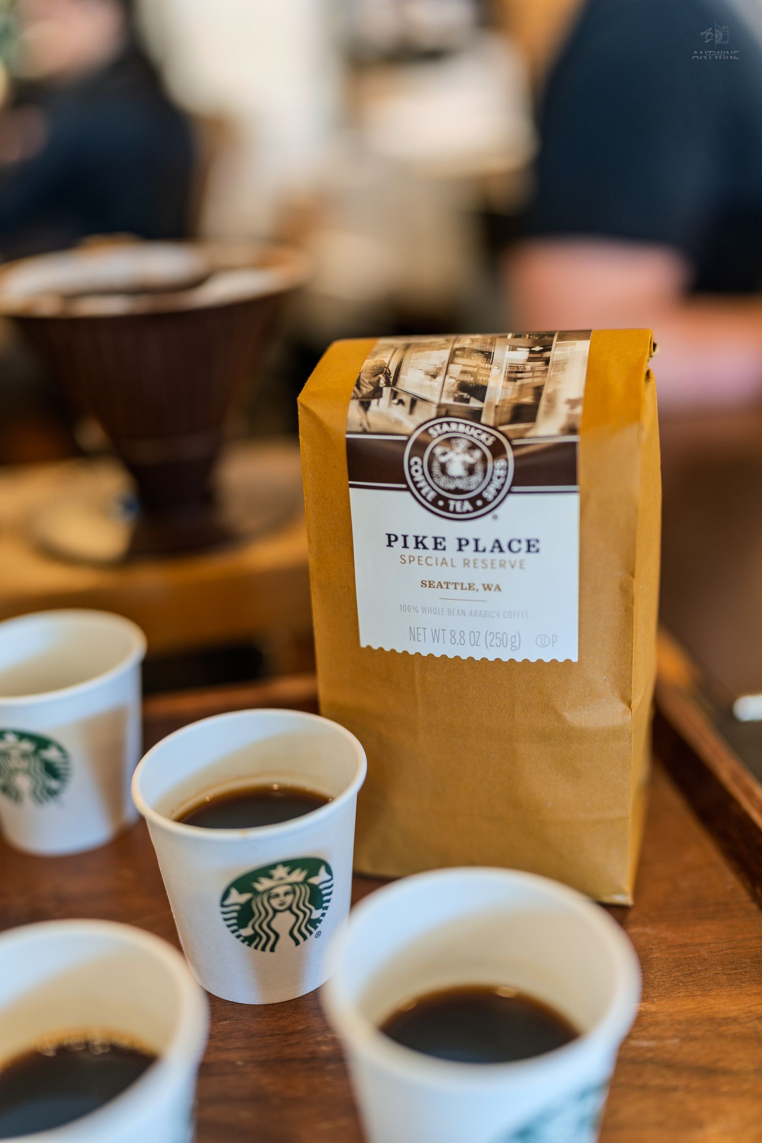 Starbucks Pike Place Special Nutmeg coffee bag surrounded by small white Starbucks cups filled with black coffee on wooden table.