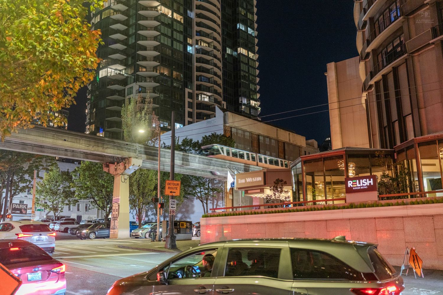 Nighttime urban street scene with illuminated skyscrapers, elevated walkway, and city traffic in a modern downtown district.