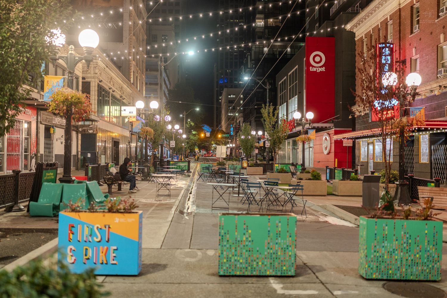 Night view of First & Pike street in Seattle with string lights, outdoor seating, and a Target store illuminated in background.