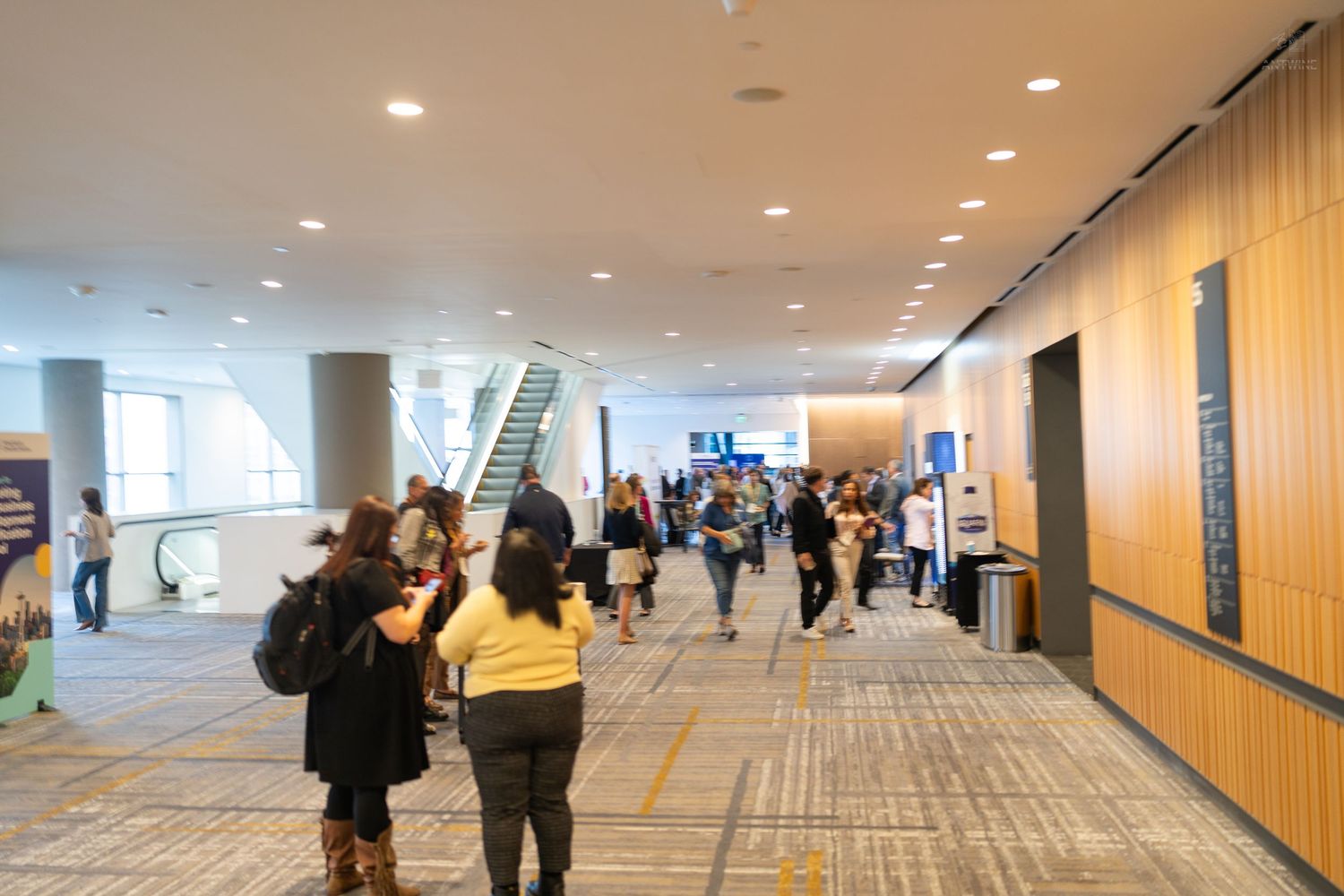 Busy convention center hallway with attendees walking past escalators and exhibit booths under bright ceiling lights.