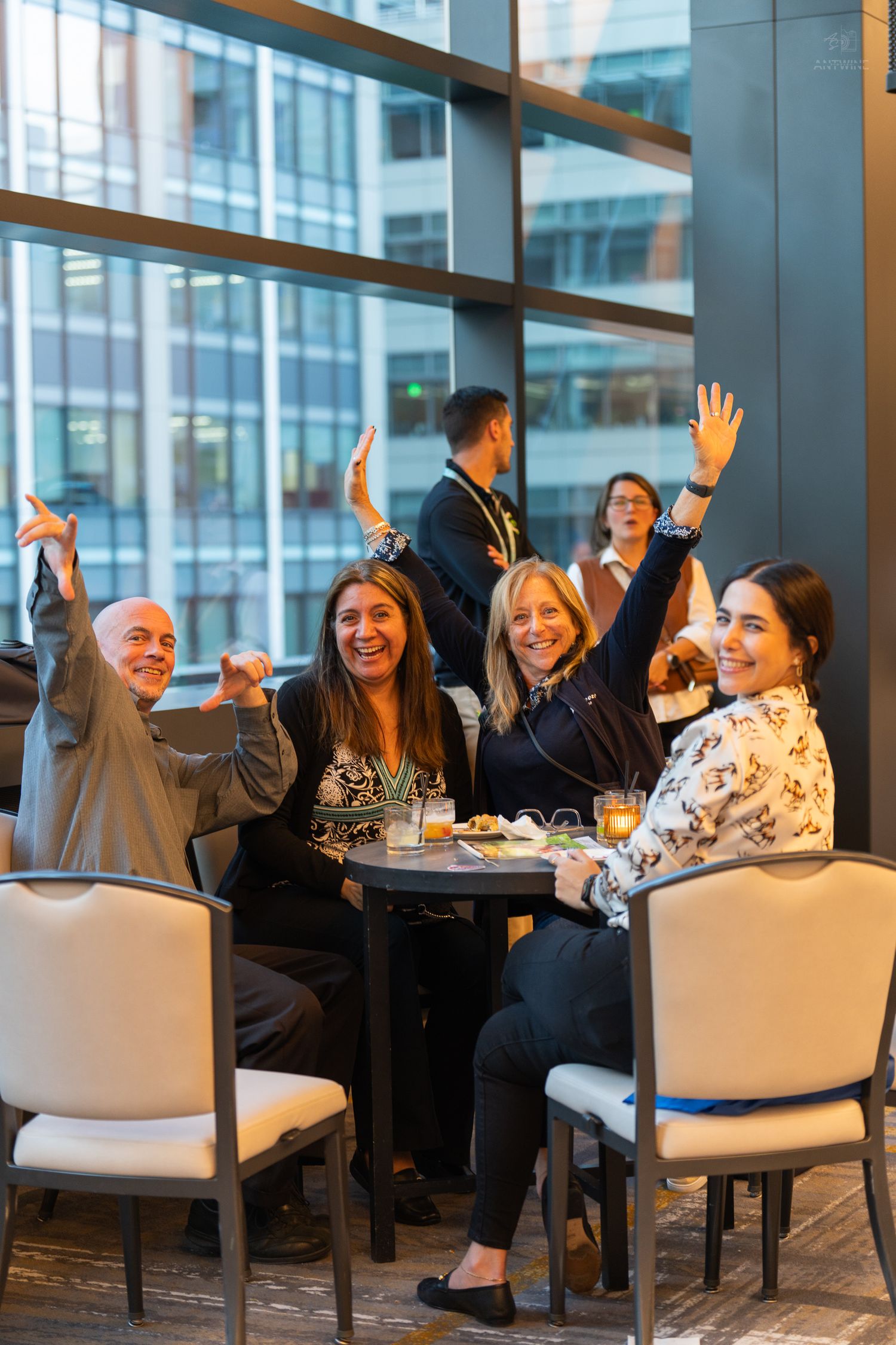 Four happy people raise their hands and smile at a table in a modern venue with large windows.
