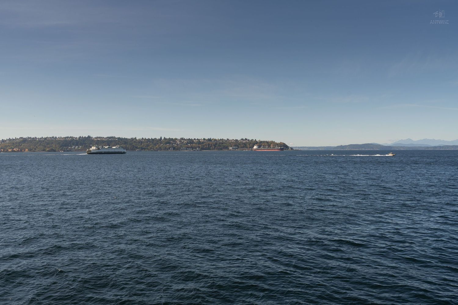 Calm blue waters of Puget Sound with ferry boat and forested shoreline under clear sky in Seattle.