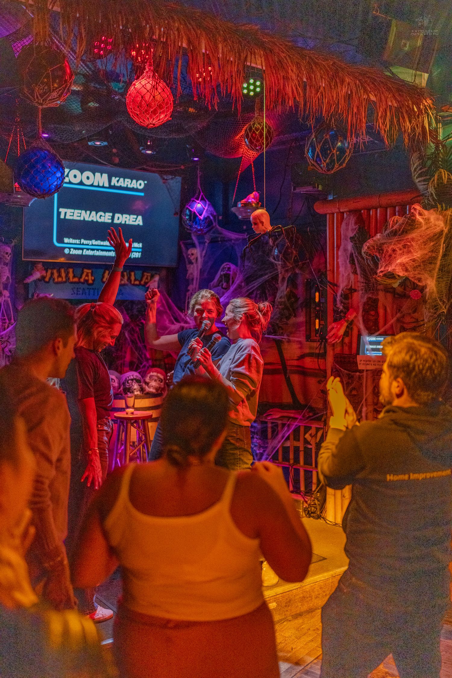 Crowd enjoying live karaoke performance at a colorfully lit tropical-themed bar at night.