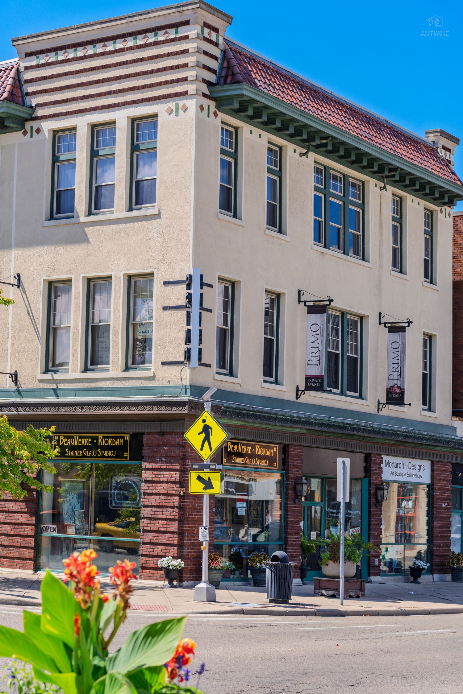 Corner commercial building with boutique shops, pedestrian crossing sign, and colorful flowers in foreground.