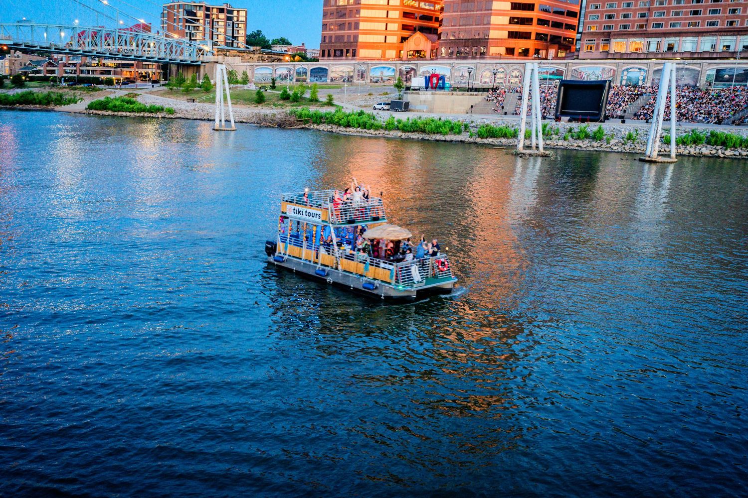 A small ferry boat carries passengers along a river with a colorful city skyline backdrop.