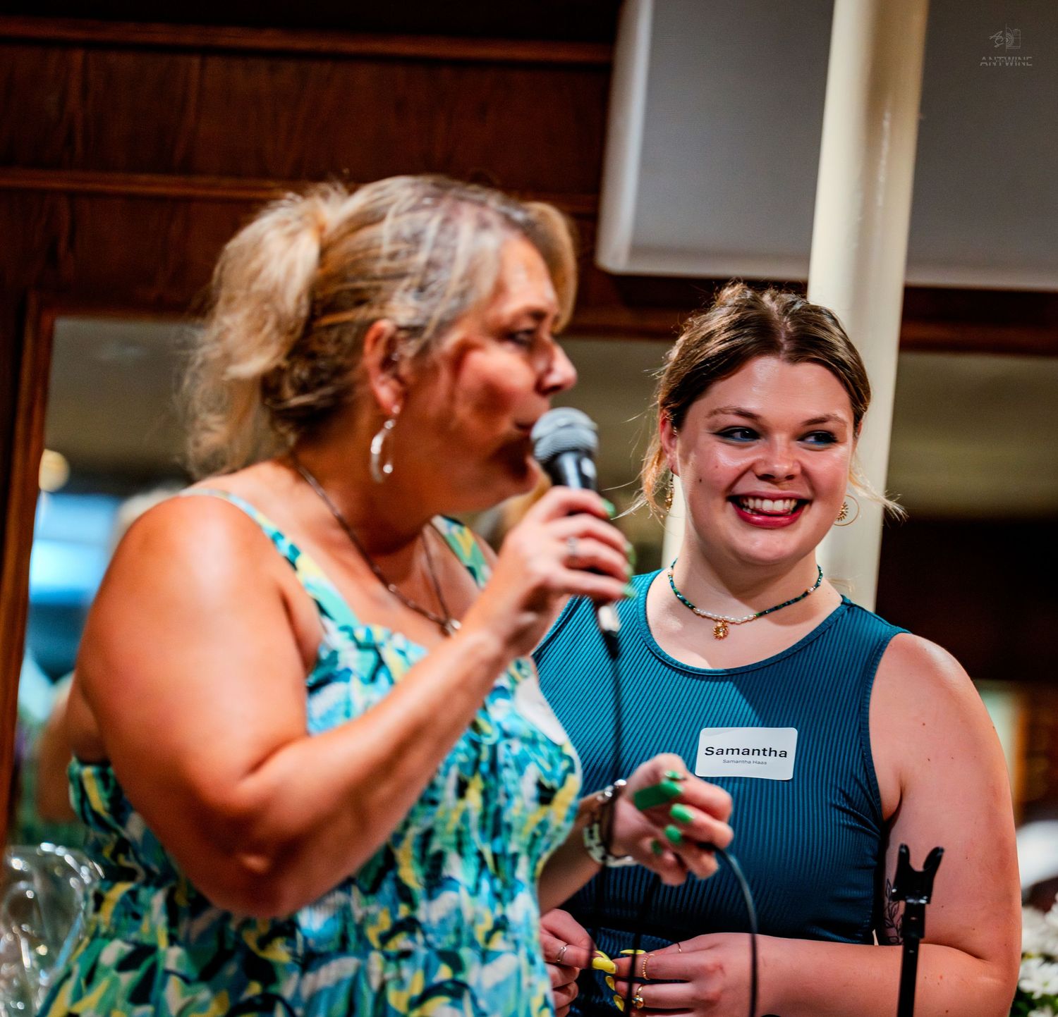 Two women smile as one speaks into a microphone at an indoor gathering or award event.