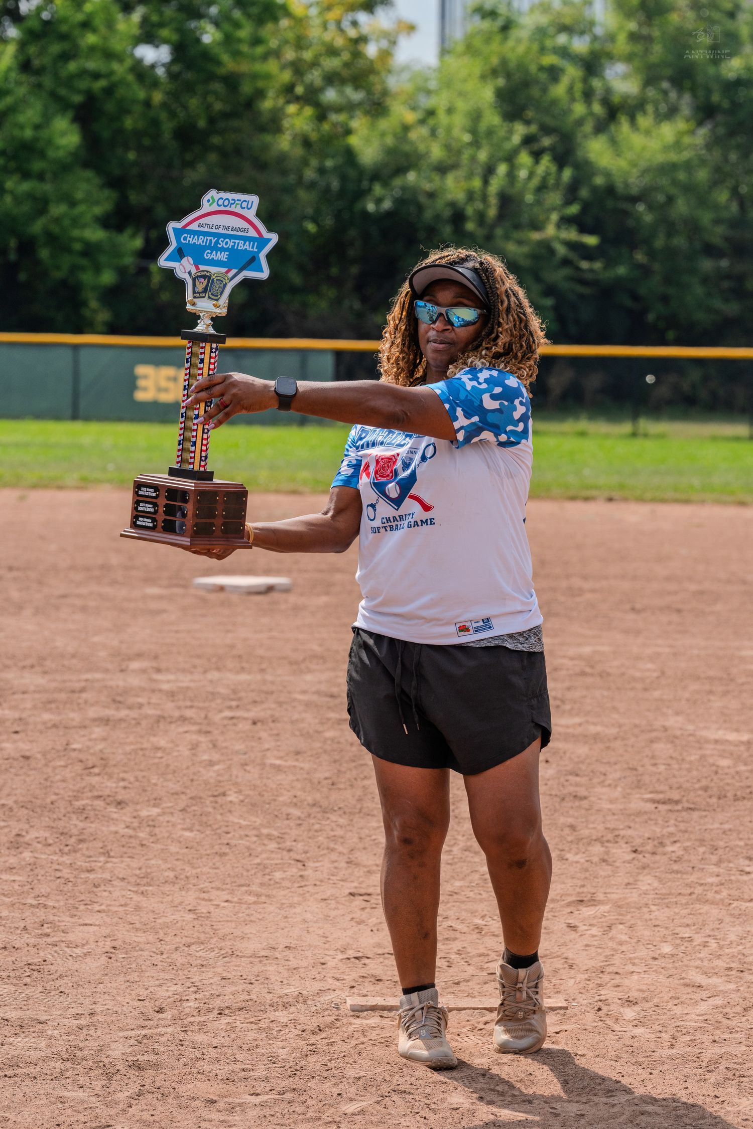 Woman proudly holds a charity softball game trophy on a baseball field on a sunny day.
