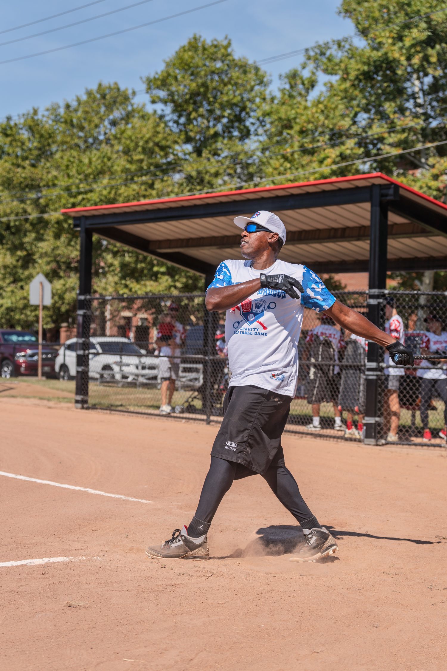 Softball player in patriotic jersey following through on swing at sunny outdoor field.