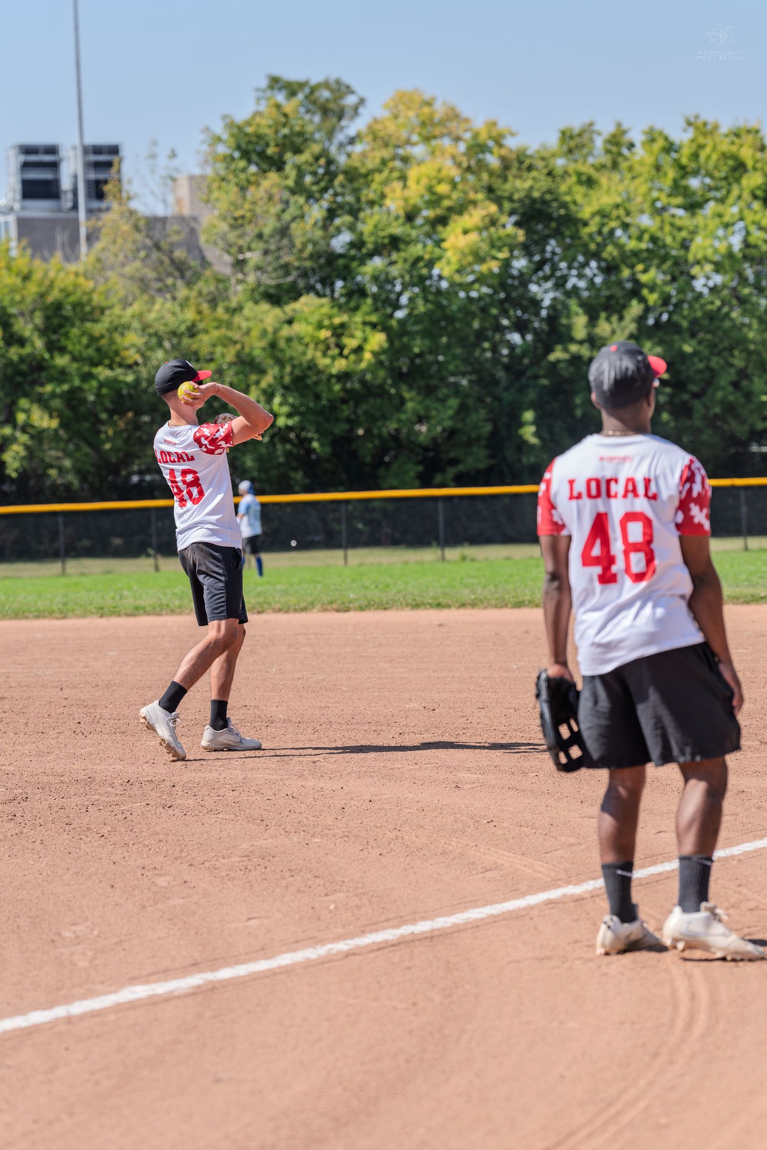 Two softball players in red Local jerseys positioning on sunny dirt infield.