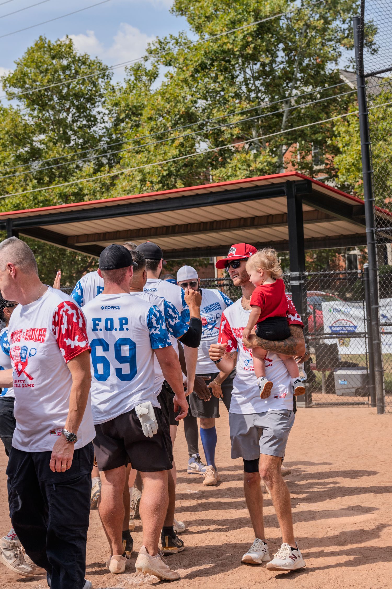 FOP softball team players in jerseys gather near dugout at outdoor baseball field on sunny day.