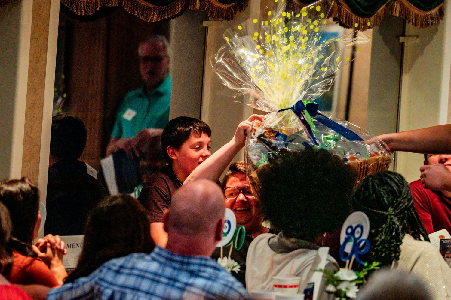 A young boy reaches for a large cellophane-wrapped gift basket at a crowded indoor charity event.