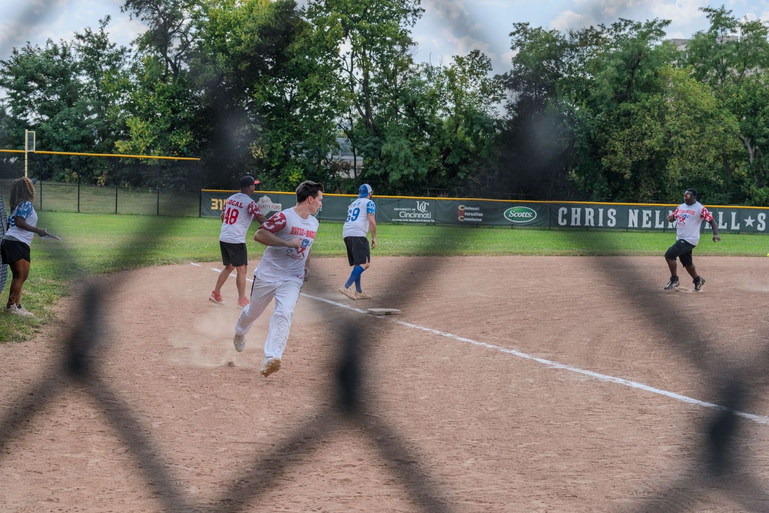 Softball players run the bases on a dirt field viewed through a chain-link fence during a game.