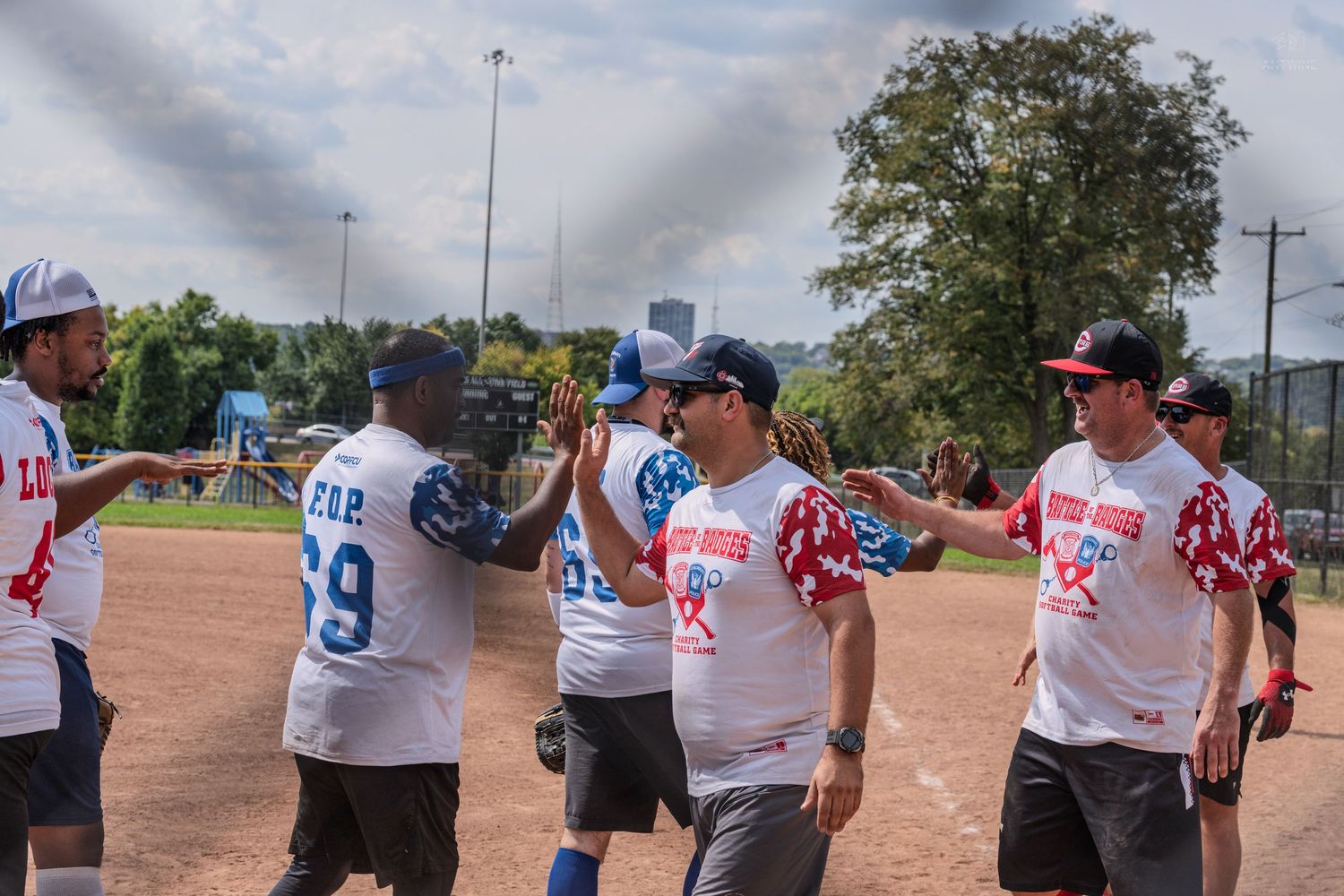 Softball players in red and blue uniforms high-fiving on a baseball diamond during a sunny outdoor game.