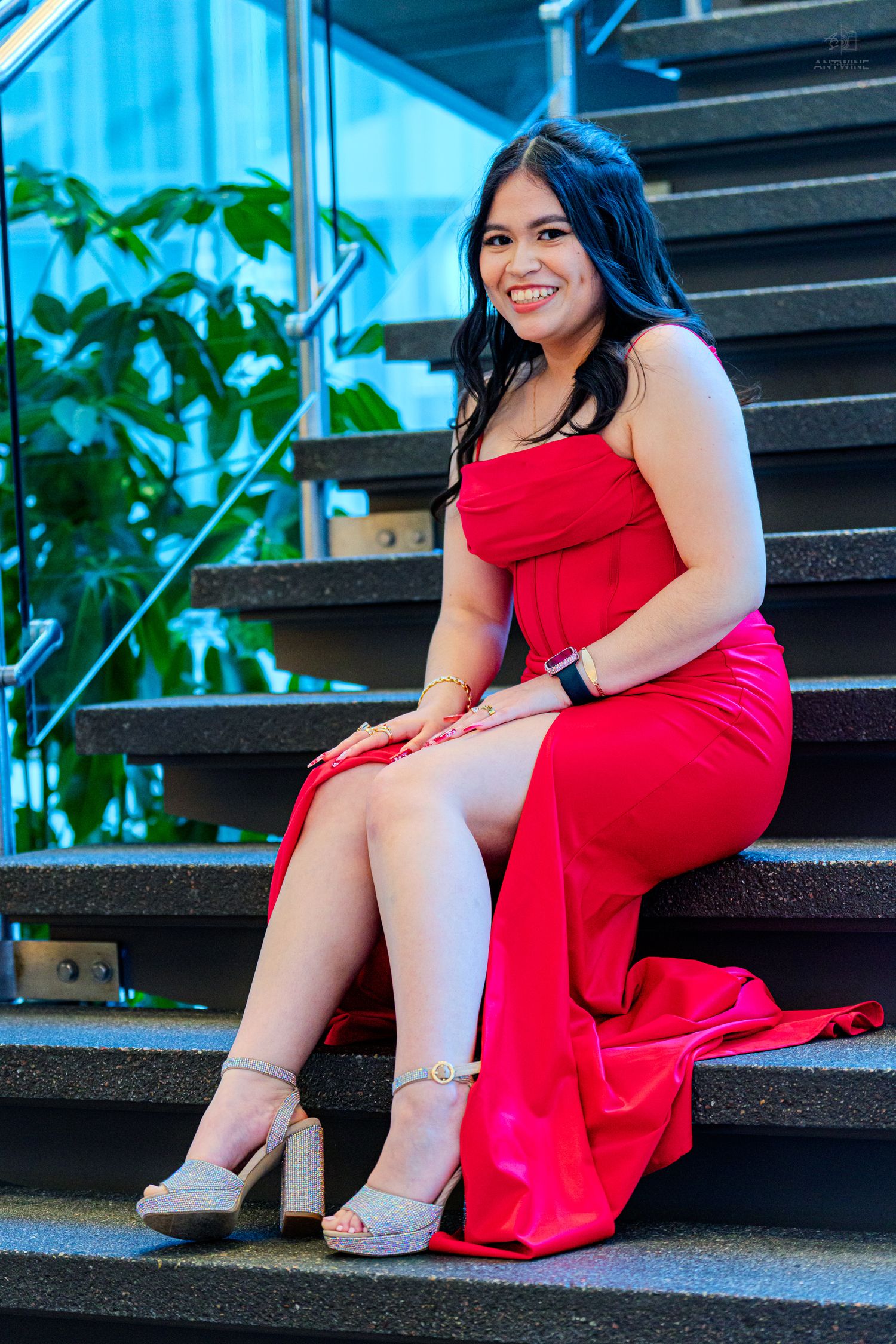 Young woman in red strapless gown and heels sitting on dark stairs, smiling outdoors.