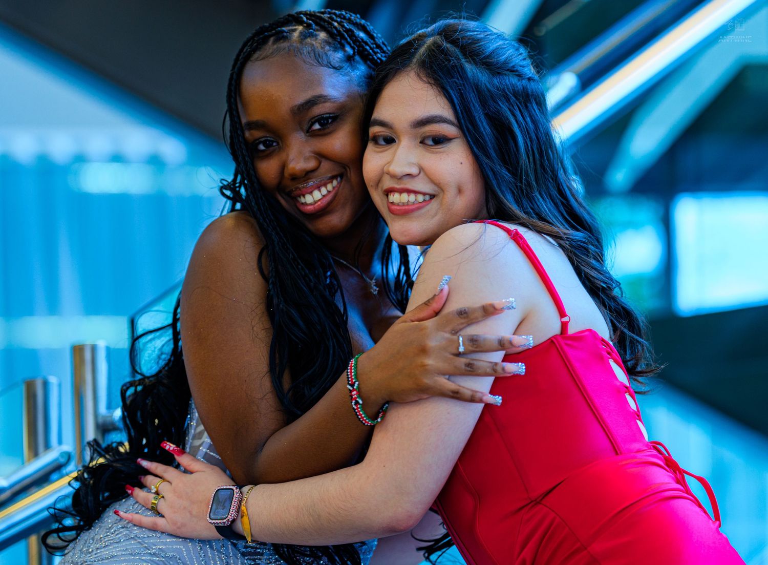 Two young women smiling and hugging each other, one in a red dress and one in a gray outfit, posing together indoors.