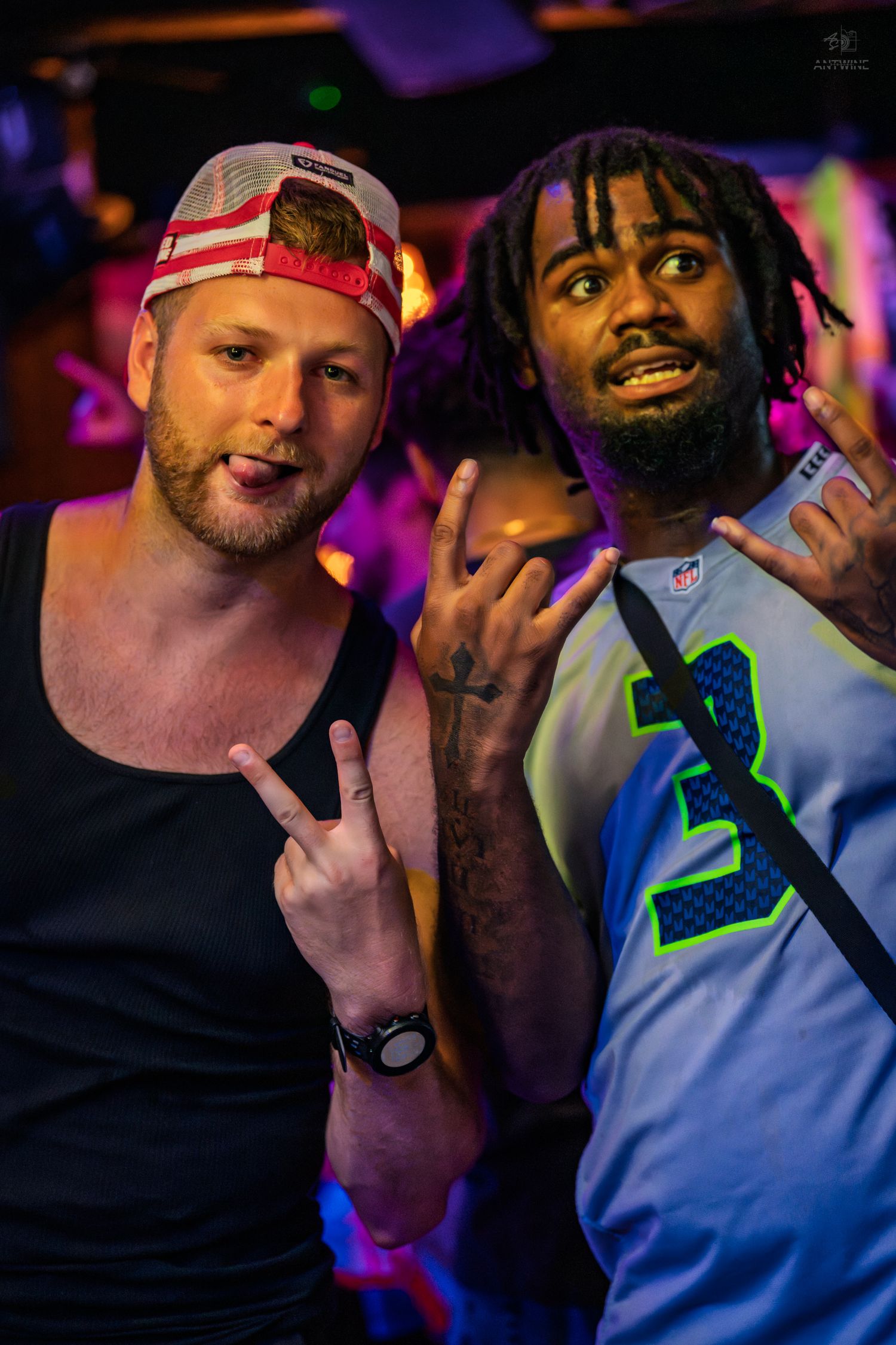 Two men smiling and posing at a nightclub, one wearing a Seahawks jersey and flag cap.