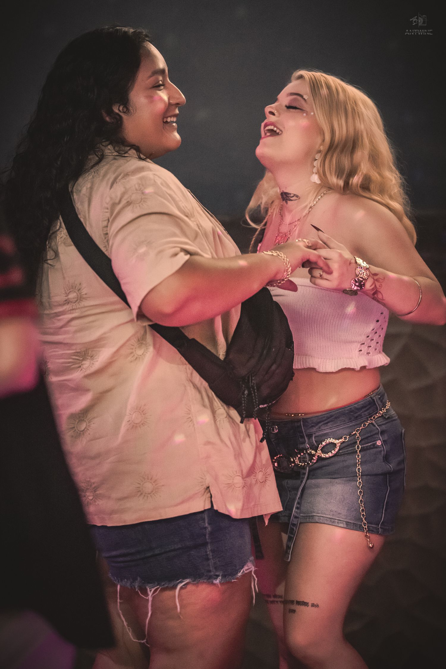 Two women laughing and dancing together at a concert, one in a white crop top and denim shorts.