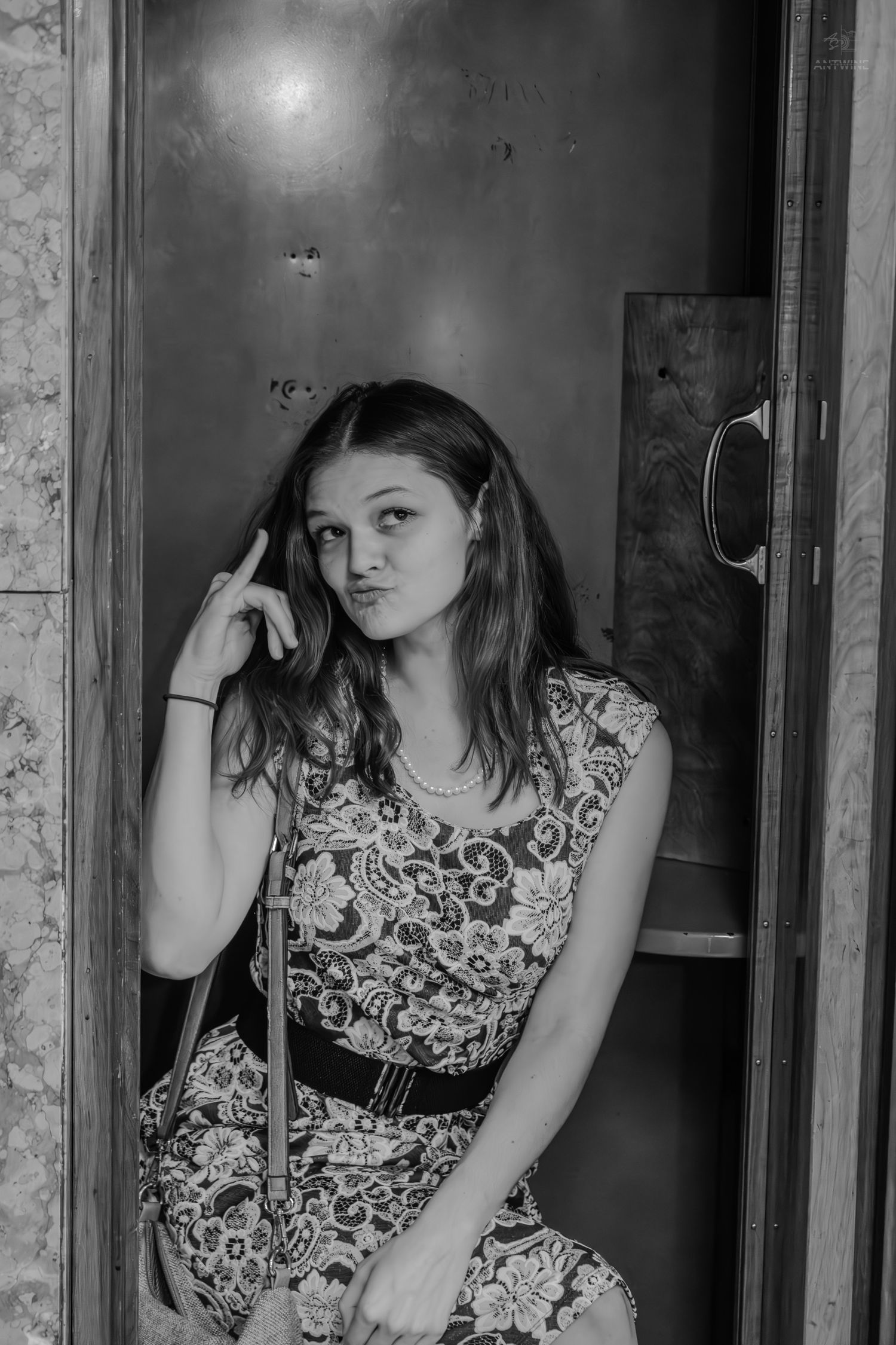 Black and white photo of a young woman in a floral dress sitting in a phone booth, looking thoughtful.