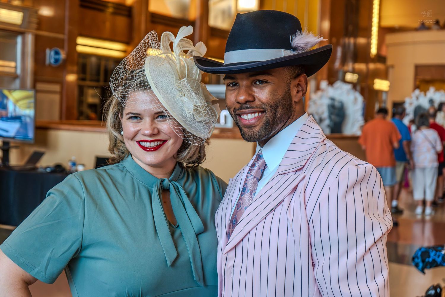 Elegantly dressed couple in vintage-style attire with fascinators and pinstripe suit at a formal event.