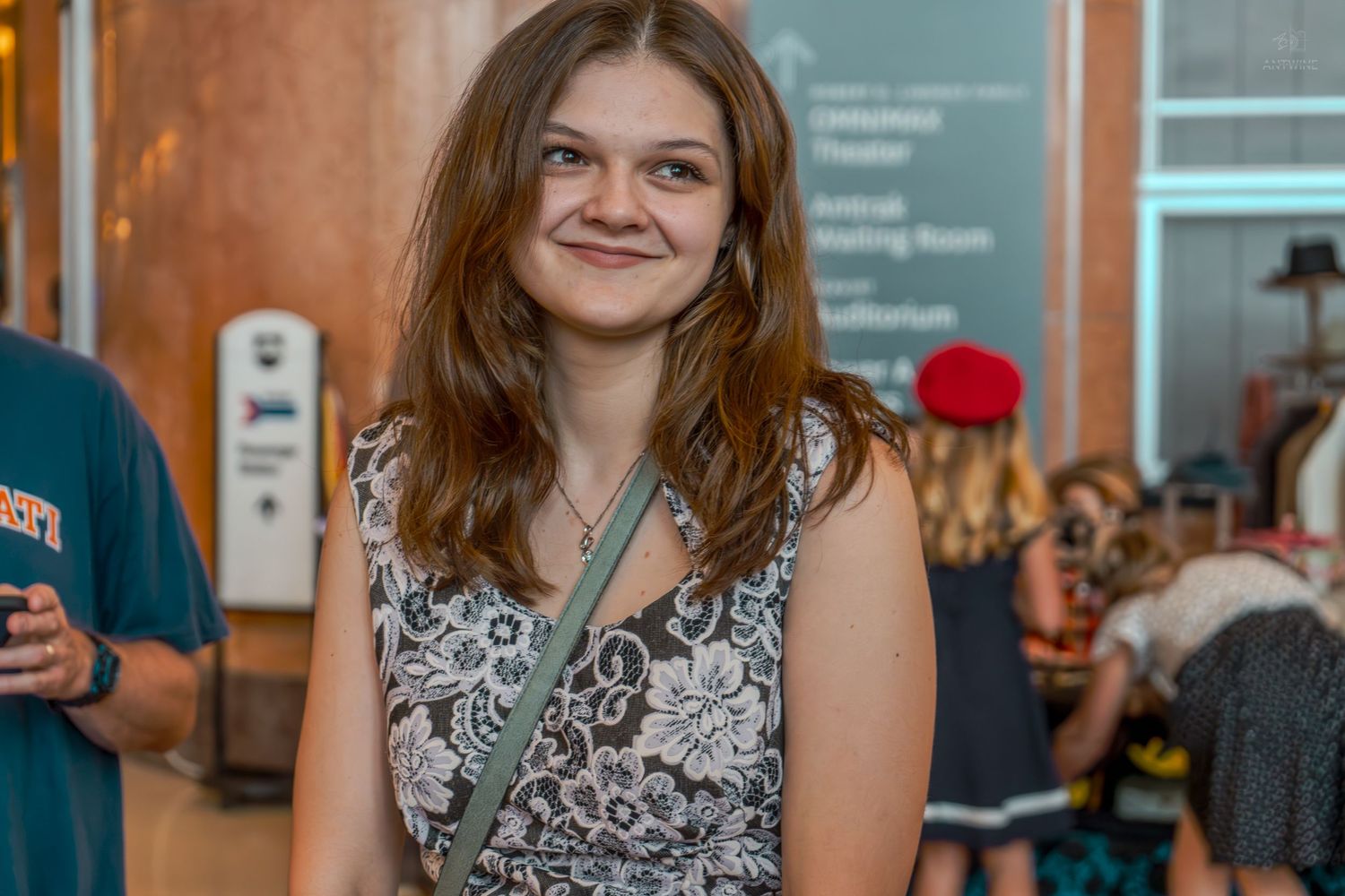 Young woman with brown hair smiling in a floral dress with a crossbody bag inside a theater lobby.