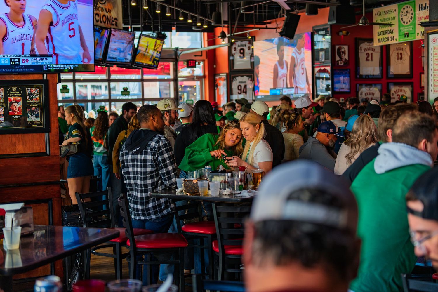 Busy sports bar packed with fans watching games on multiple TVs, enjoying drinks at red-chaired tables.