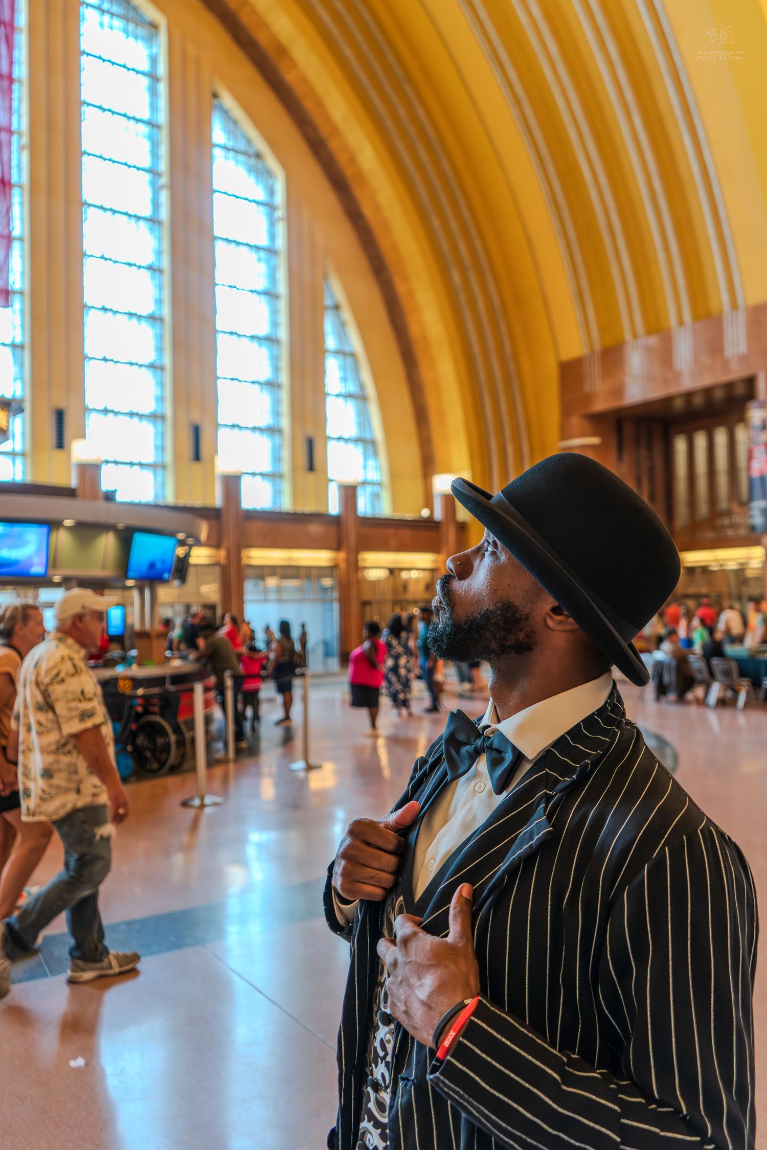 Stylish Black man in pinstripe suit and bowler hat poses in grand train station hall.