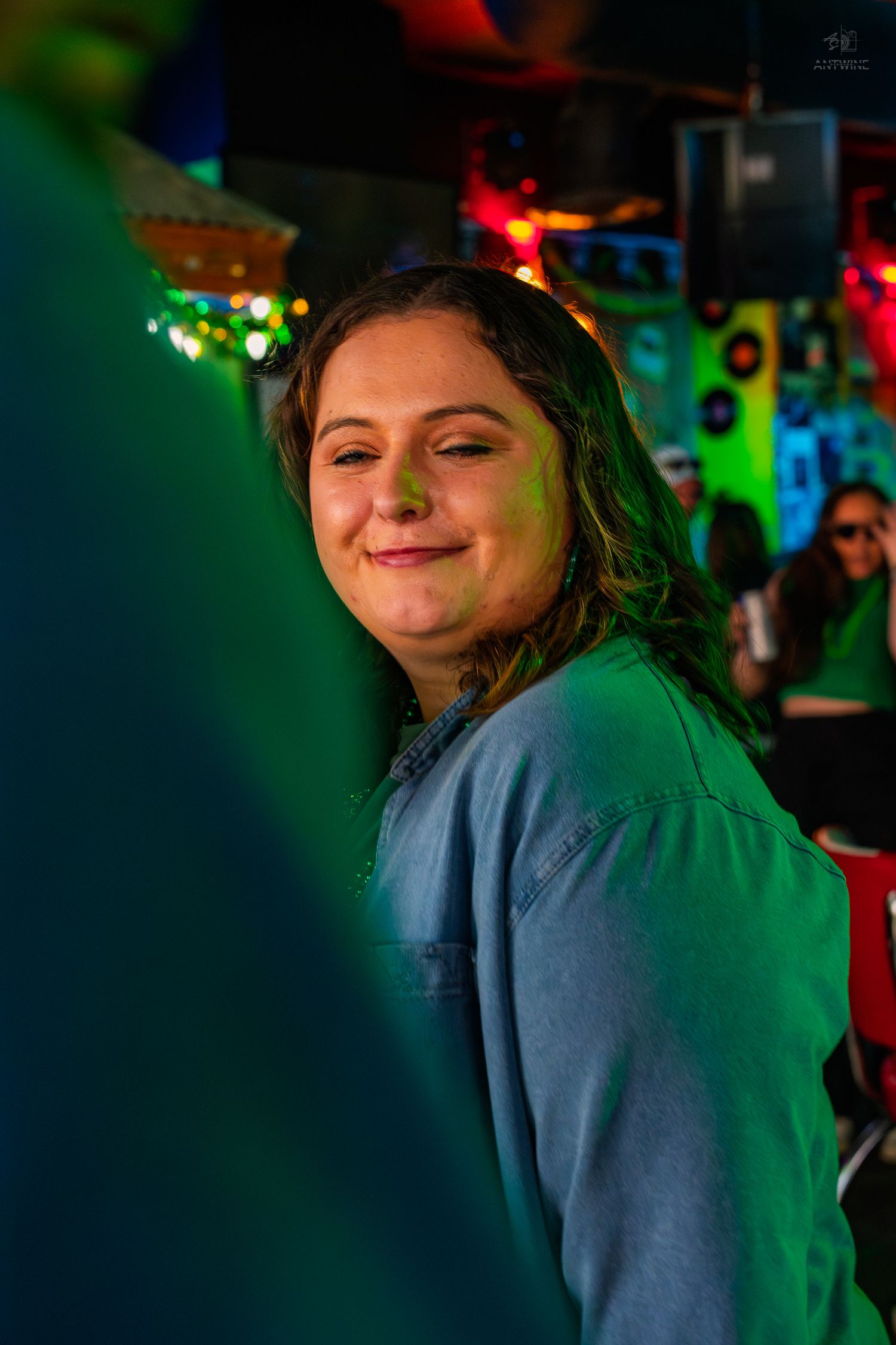A smiling young woman in a denim shirt at a vibrant, colorfully lit bar or club with green and red lights.