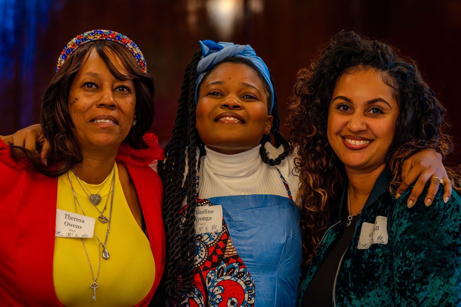 Three women smiling and posing together at an indoor event, wearing name badges and colorful outfits.