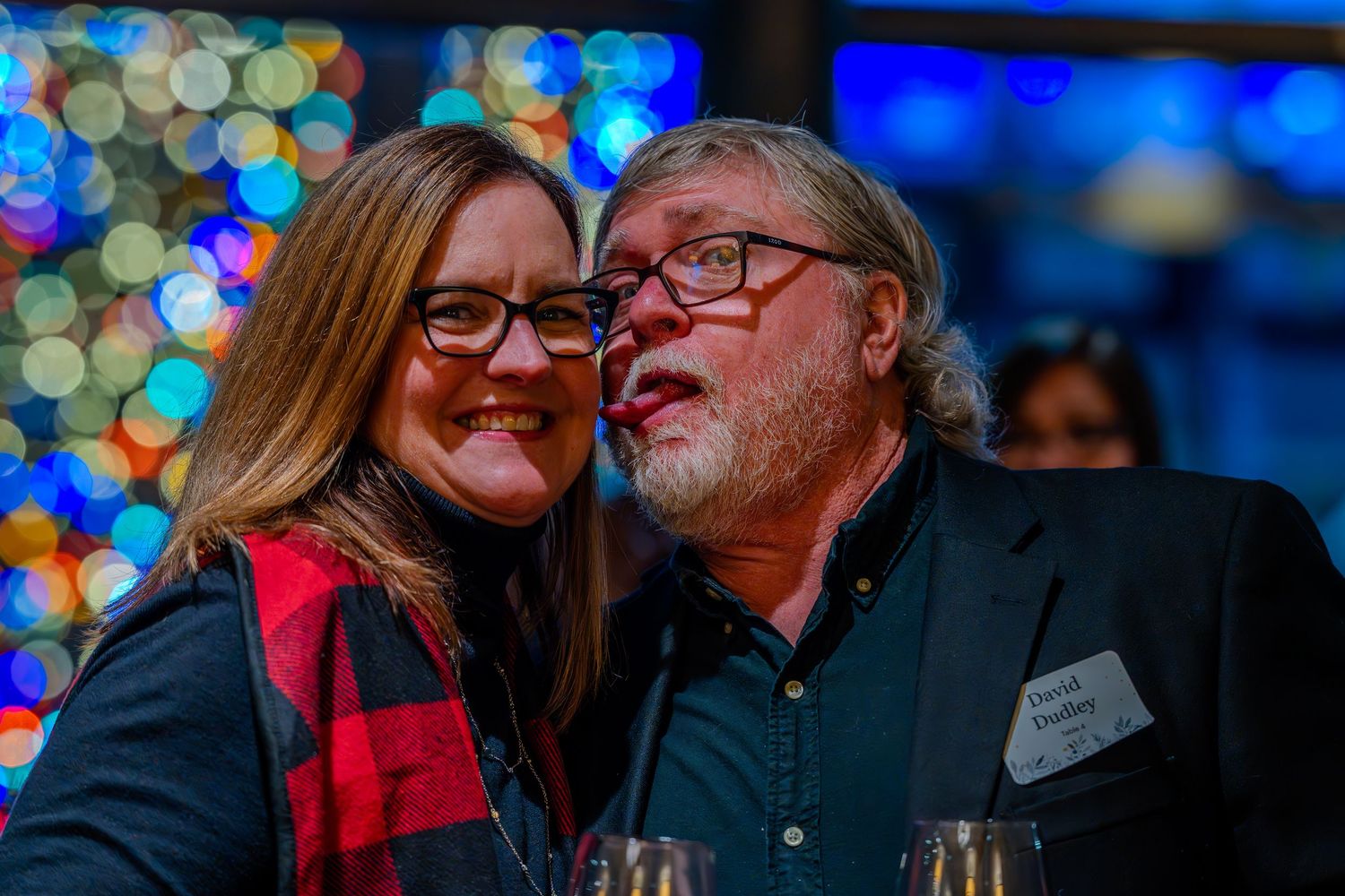 Two smiling adults pose closely together at a festive holiday party with colorful Christmas tree lights in background.