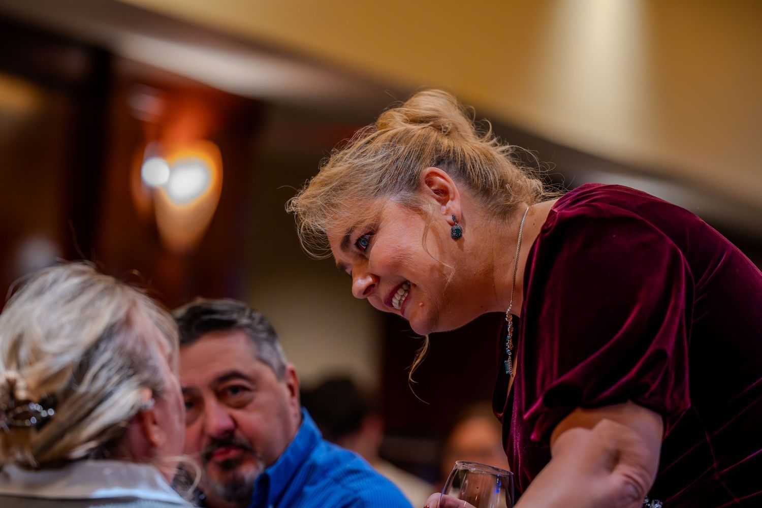 Woman in burgundy velvet top smiling and leaning toward seated guests at an indoor dining event.