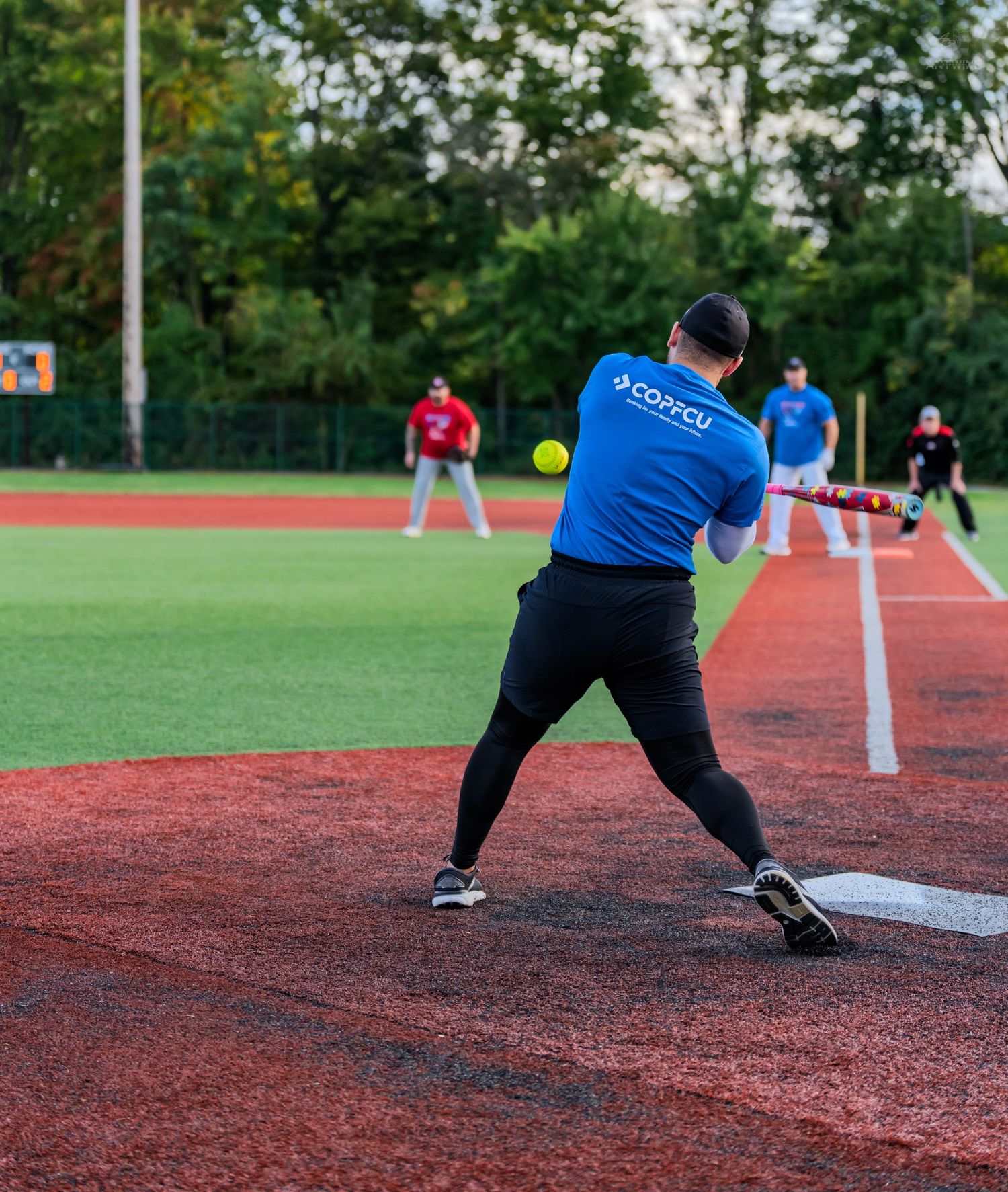 Batter in blue shirt swinging at yellow softball during an outdoor game on red turf field.