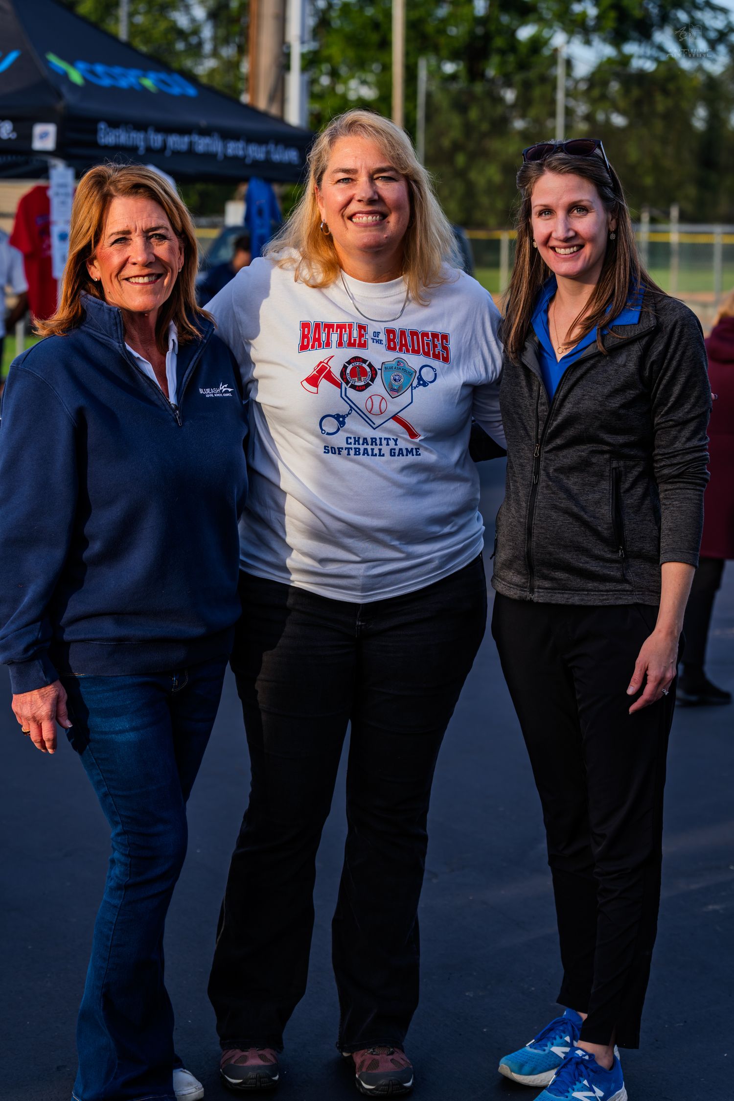 Three women smile together at a Battle of the Badges charity softball game event outdoors.