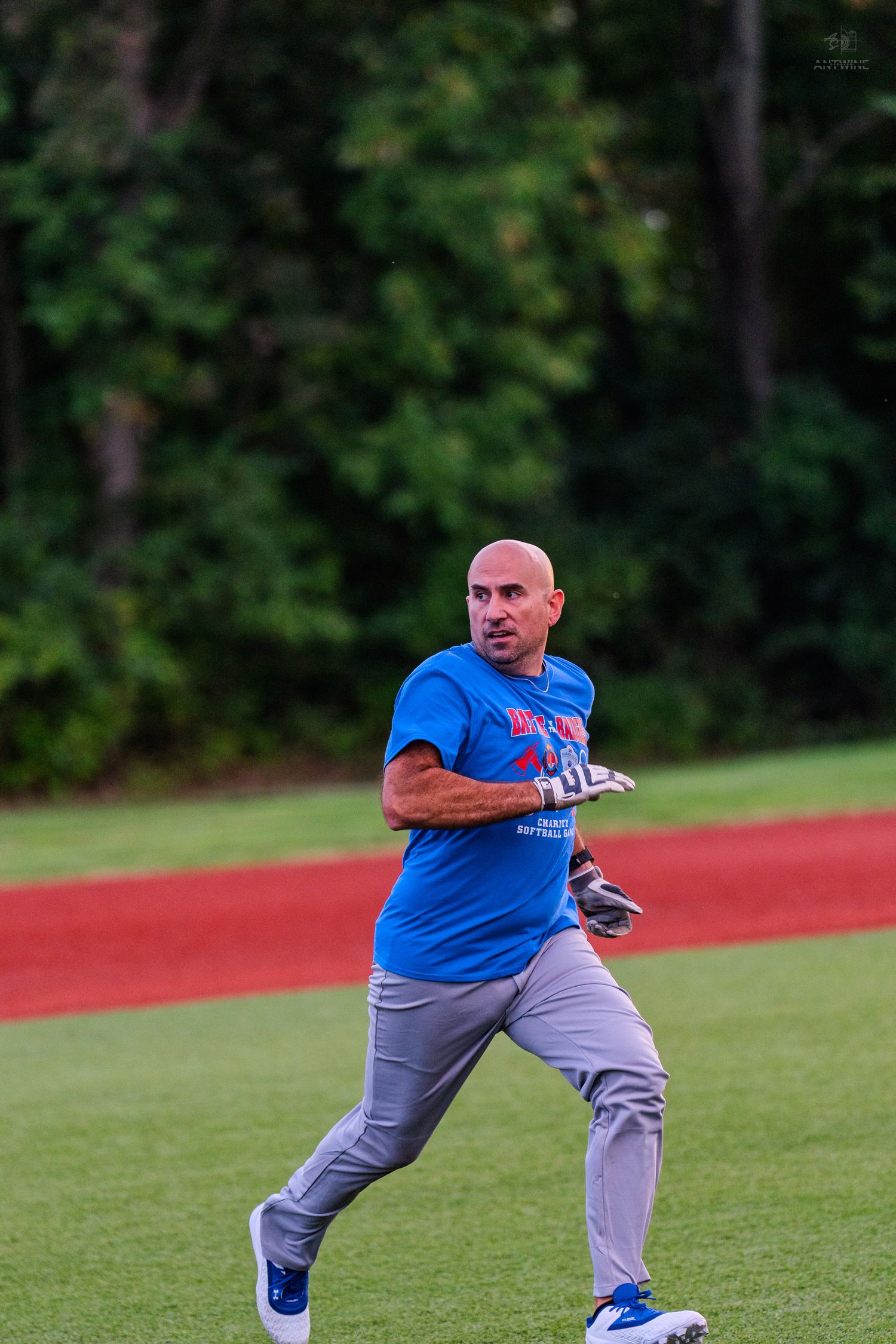 Bald man in blue t-shirt playing softball, mid-throw on a green field with red warning track.