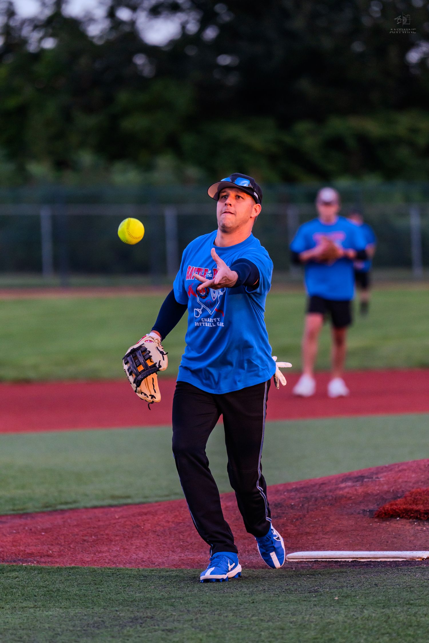 Softball player in blue jersey throws yellow ball during evening recreational league game.