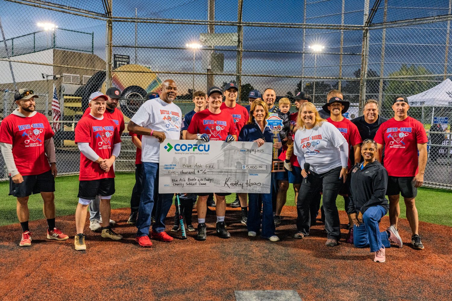 Group of people holding a large CO-FCU charity check at a baseball field fundraiser event at night.