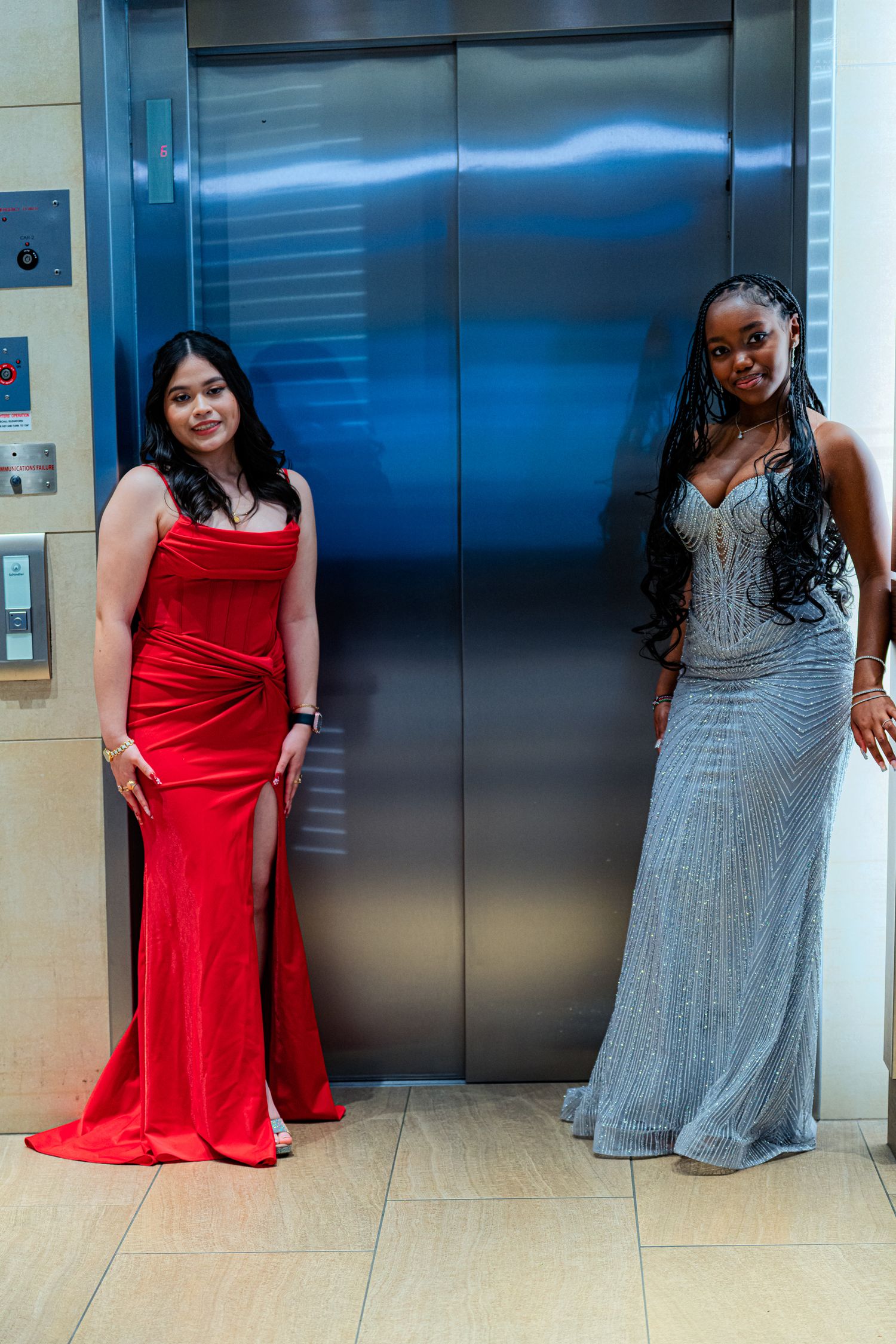 Two young women in elegant prom gowns, one red and one silver, posing in front of a shiny elevator.