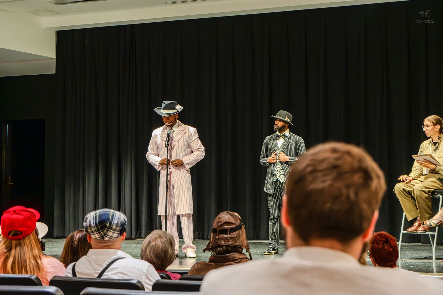 Two performers in vintage suits and hats on stage before a seated audience in a black curtain theater setting.