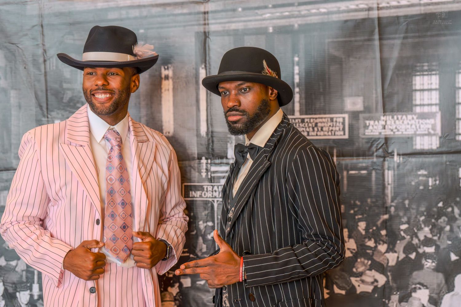Two men in vintage pinstripe suits and fedora hats pose elegantly in front of a historic black and white photograph.
